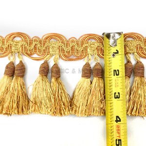 May include: Gold and brown tassel trim with a wavy design. The trim is measured with a tape measure showing it is 2 and 1/2 inches wide.
