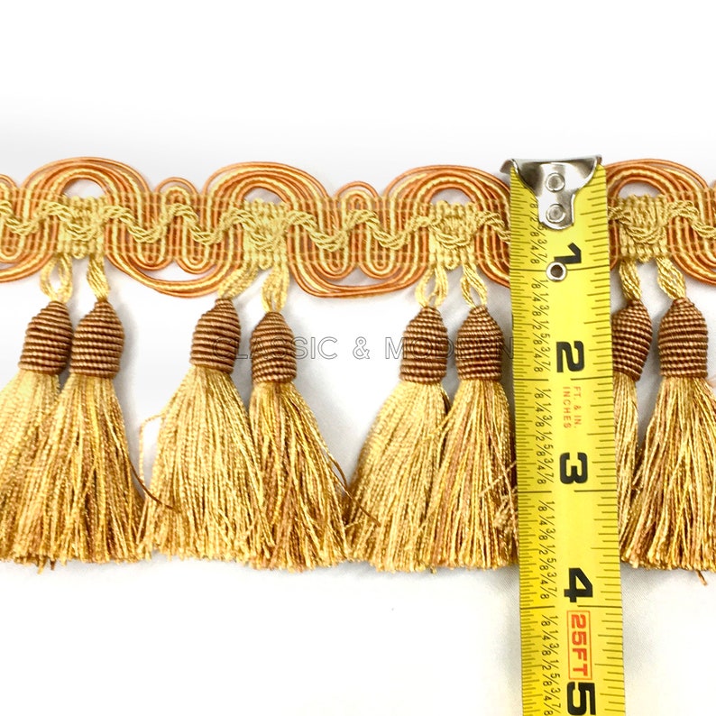 May include: Gold and brown tassel trim with a wavy design. The trim is measured with a tape measure showing it is 2 and 1/2 inches wide.