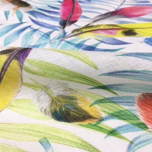 May include: Close-up of a white fabric with a colorful tropical print featuring green leaves, blue palm fronds, and yellow, red, and blue feathers.