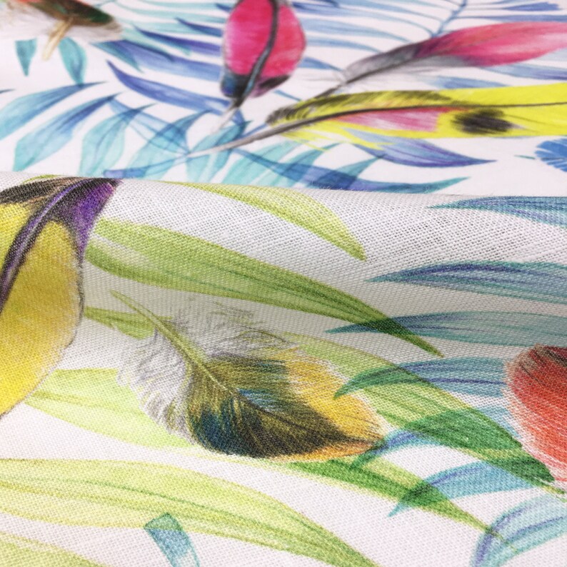 May include: Close-up of a white fabric with a colorful tropical print featuring green leaves, blue palm fronds, and yellow, red, and blue feathers.