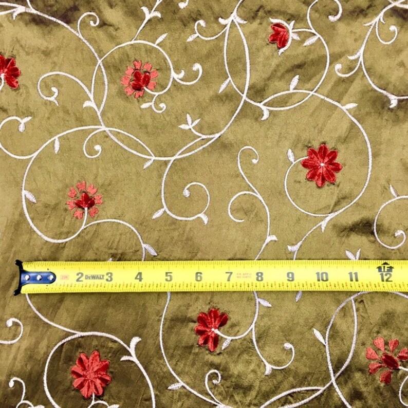 May include: A close-up of a brown fabric with a white floral pattern and red embroidered flowers. A yellow measuring tape is placed across the fabric, showing measurements in inches.