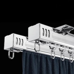 Può includere: Primo piano di un sistema di bastone per tende color argento con una tenda blu scuro. Il bastone ha una guida con più ganci per appendere le tende. Le estremità sono quadrate con ritagli decorativi.