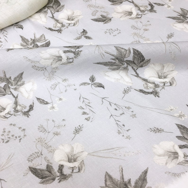 May include: A light gray fabric with a white floral pattern. The flowers are stylized and have a delicate, watercolor-like appearance. The fabric has a subtle texture.