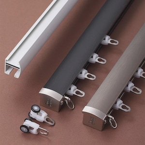 Può includere: Primo piano di tre bastoni per tende in bianco, grigio scuro e grigio chiaro. I bastoni hanno una forma rettangolare con una guida per i ganci delle tende. Due rulli per tende bianchi sono in primo piano. I bastoni hanno terminali metallici.