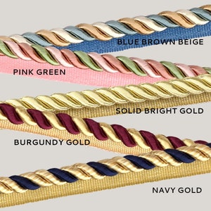 May include: Close-up of five different types of gold trim rope with various colors, including blue, brown, beige, pink, green, burgundy, navy, and solid bright gold. The trim rope is a decorative element for home decor and crafts.
