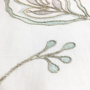 May include: Close-up of a white fabric with embroidered light green and brown floral designs. The design features a branch with three leaves.