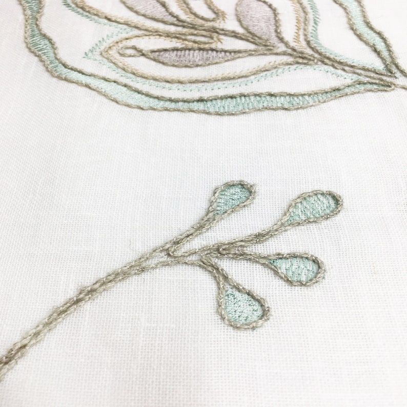 May include: Close-up of a white fabric with embroidered light green and brown floral designs. The design features a branch with three leaves.