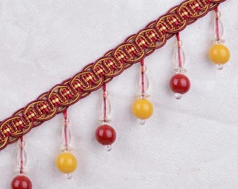 May include: Decorative trim featuring a burgundy and gold braided design. The trim has alternating red and yellow beaded accents suspended from the braid. The trim is likely used for home decor or craft projects.
