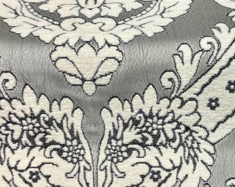May include: Close-up of a fabric with a damask pattern. The design features white floral motifs outlined in black against a gray background. The fabric has a textured appearance, suggesting a woven material.