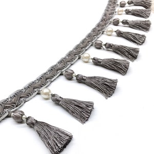 May include: Grey trim with a woven design and pearl and grey bead accents. The trim has a fringe of grey tassels.