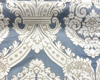 May include: Close-up of a blue fabric with a damask pattern in white and gold. The fabric has a soft sheen and a detailed, repeating design of swirling leaves and floral elements. The pattern is symmetrical and ornate.