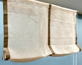 May include: White roman blind with brown trim. The blind is made of a woven fabric and has a cord to raise and lower it.
