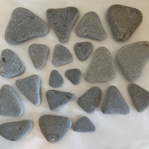 May include: An assortment of gray, smooth beach stones in various shapes, including triangles and rounded forms. The stones are arranged on a white surface, showcasing their natural textures and colors. Ideal for crafts or decorative purposes.