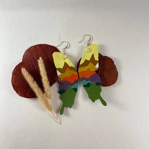 Puede incluir: Un par de pendientes de mariposa coloridos con un diseño de montaña en capas. Los pendientes están hechos de madera pintada y tienen ganchos plateados.