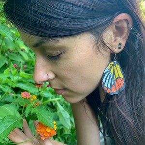 Puede incluir: Una mujer con el pelo largo y oscuro huele un racimo de flores naranja y amarillas. Lleva un par de pendientes con forma de mariposa con detalles en negro, blanco, amarillo y naranja.