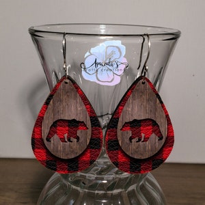 May include: Red and black plaid teardrop shaped earrings with a woodgrain background and a black bear silhouette.
