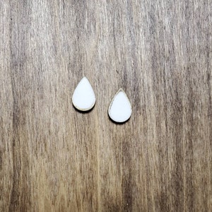 以下が含まれることがあります： 茶色の木製の表面に置かれた、白い涙の形をしたイヤリングのペア