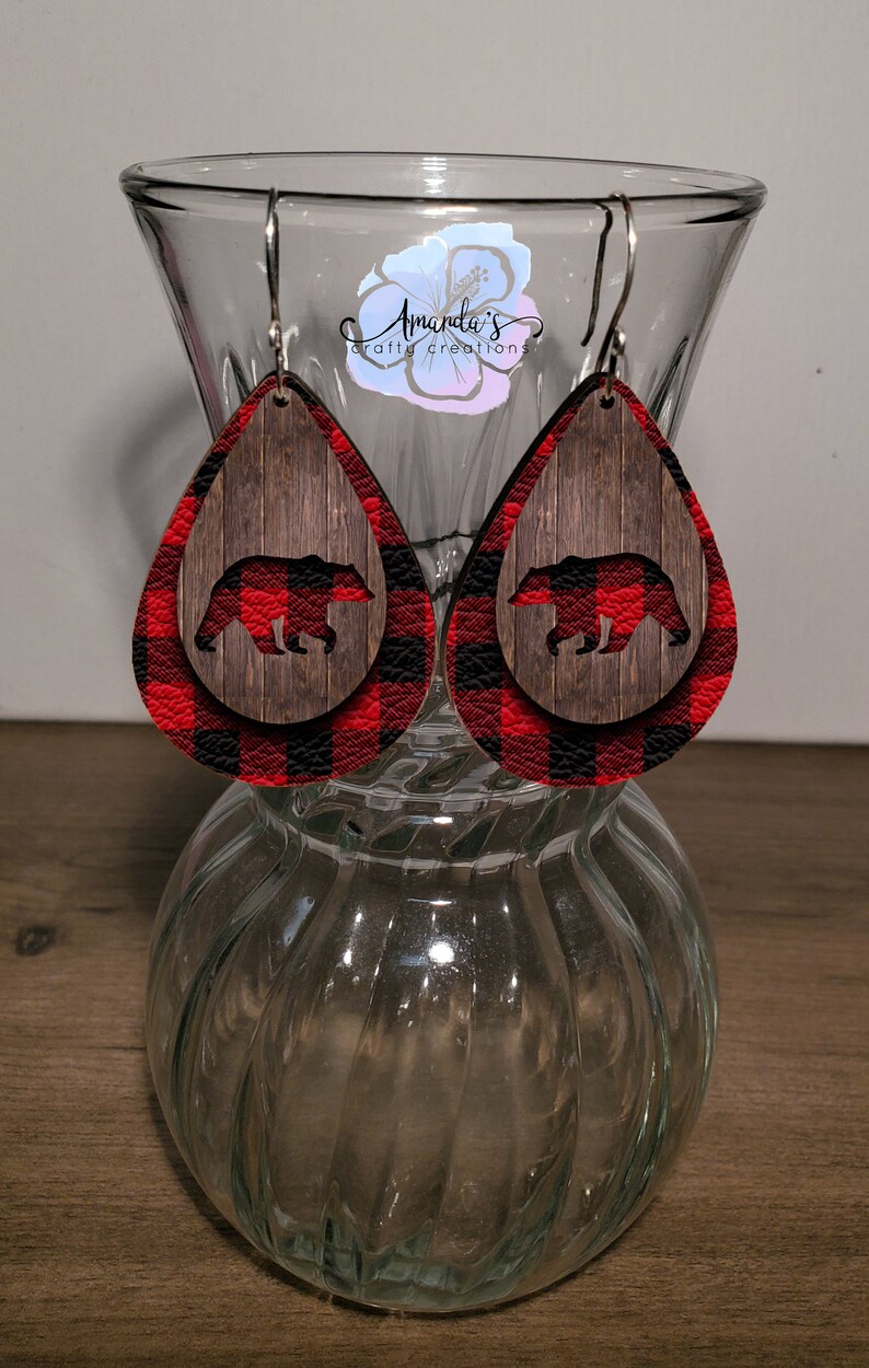 May include: Red and black plaid teardrop shaped earrings with a woodgrain background and a black bear silhouette.