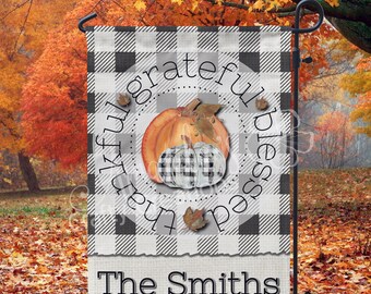 May include: A black and white checkered garden flag with a circle in the center that says "Thankful Grateful Blessed" with a pumpkin and fall leaves. The flag has a white banner at the bottom that says "The Smiths".