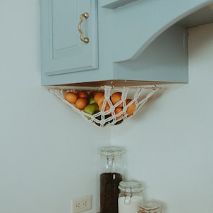 May include: A white string hammock hanging from a light blue cabinet, filled with oranges and limes. Three glass jars with lids sit on a wooden countertop below the hammock.