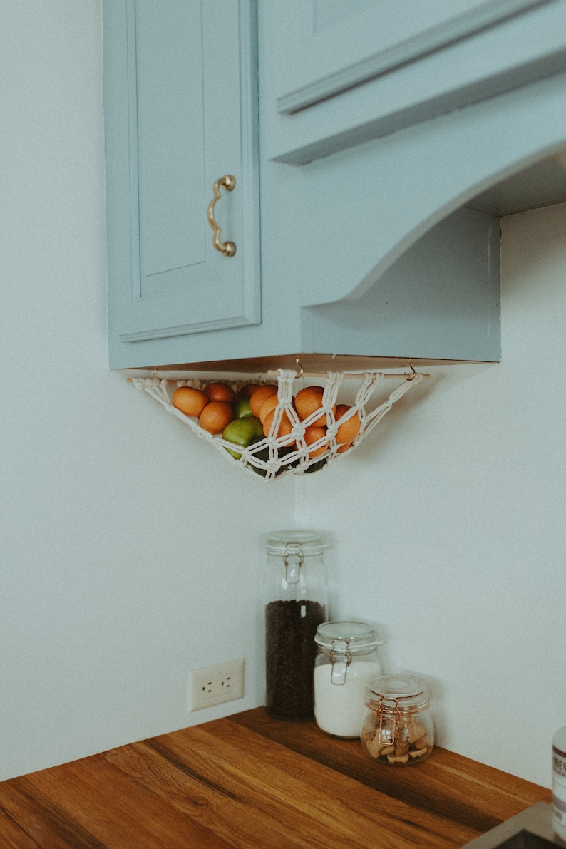 May include: A white string hammock hanging from a light blue cabinet, filled with oranges and limes. Three glass jars with lids sit on a wooden countertop below the hammock.