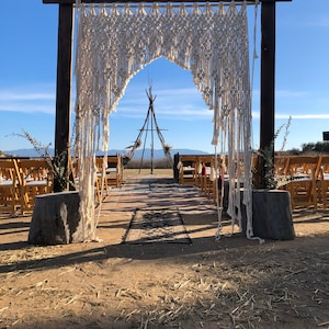 May include: A rustic wooden archway decorated with a white macrame hanging. The archway is set up for an outdoor wedding ceremony with chairs lined up in rows leading to the archway.