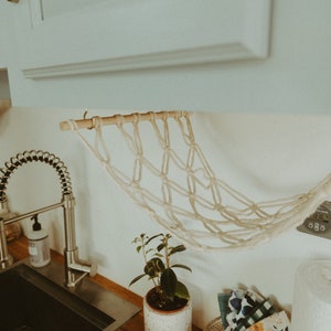 May include: A white macrame hanging fruit basket with a wooden dowel rod, hanging from a white cabinet in a kitchen. The basket is hanging over a stainless steel kitchen sink with a pull-down faucet.