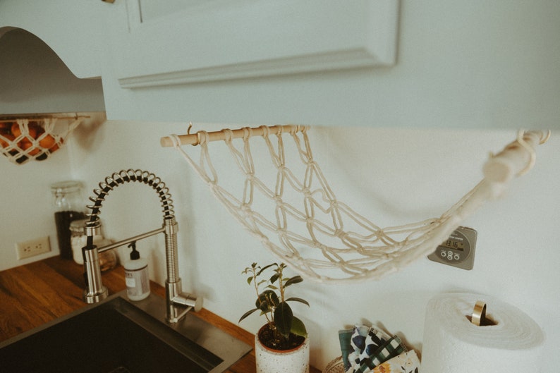 May include: A white macrame hanging fruit basket with a wooden dowel rod, hanging from a white cabinet in a kitchen. The basket is hanging over a stainless steel kitchen sink with a pull-down faucet.