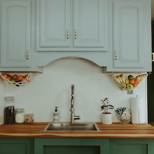 May include: A kitchen sink with a stainless steel basin and a chrome faucet. The sink is set into a wooden countertop with green cabinets below. There are two hanging fruit baskets above the sink, one with oranges and the other with bananas, apples, and oranges.