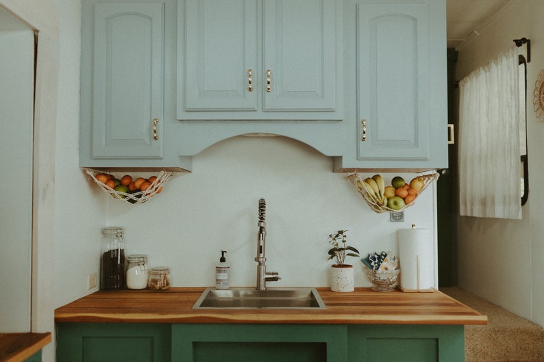 May include: A kitchen sink with a stainless steel basin and a chrome faucet. The sink is set into a wooden countertop with green cabinets below. There are two hanging fruit baskets above the sink, one with oranges and the other with bananas, apples, and oranges.