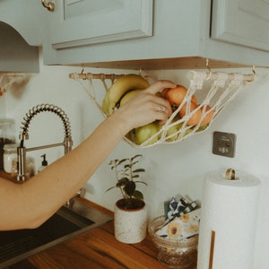 May include: A white macrame hanging fruit basket with bananas, apples, and oranges hanging from the underside of a gray kitchen cabinet. The basket is attached to the cabinet with two gold hooks.