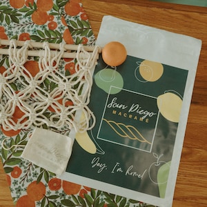 May include: A white macrame wall hanging with a wooden dowel, a small white pouch, and a printed card with the text "San Diego Macrame" and "Day, I'm home!" on a background of green, yellow, and orange circles.