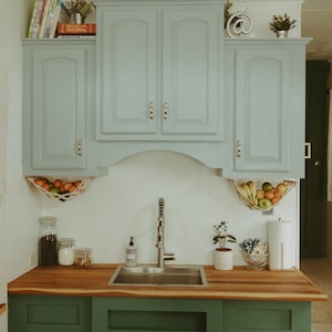 May include: A kitchen with light blue cabinets, a stainless steel sink, and green cabinets below. The cabinets have gold handles. There are fruit hanging in a net above the sink.