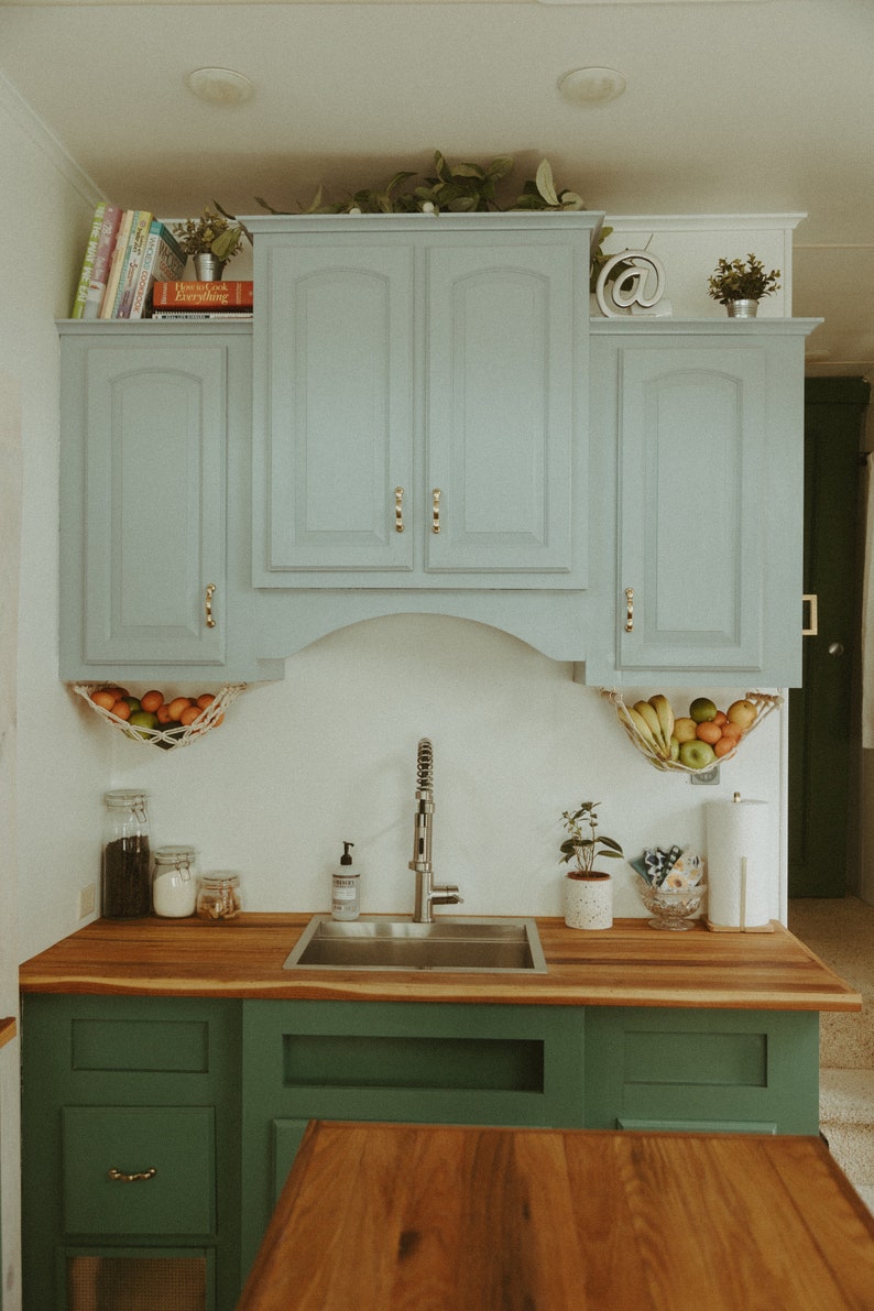 May include: A kitchen with light blue cabinets, a stainless steel sink, and green cabinets below. The cabinets have gold handles. There are fruit hanging in a net above the sink.