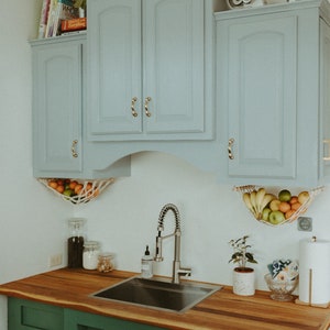 May include: A kitchen with green cabinets and a wood countertop. The cabinets have gold handles. There is a stainless steel sink and faucet. There are two hanging fruit baskets with fruit inside. There is a plant on the counter and a sign with an at symbol on the top cabinet.