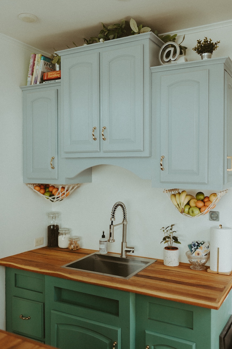 May include: A kitchen with green cabinets and a wood countertop. The cabinets have gold handles. There is a stainless steel sink and faucet. There are two hanging fruit baskets with fruit inside. There is a plant on the counter and a sign with an at symbol on the top cabinet.