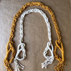 May include: Two macrame plant hangers, one in white and one in mustard yellow, with tassels at the bottom.