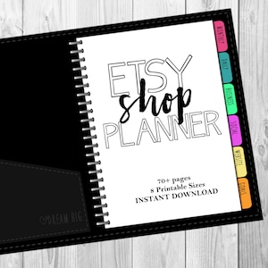 Puede incluir: Un planificador en blanco y negro con el texto "ETSY shop PLANNER" en letras negritas y con contorno. El planificador tiene una cubierta negra con una encuadernación en espiral plateada y un bolsillo negro en la parte delantera. El texto "DREAM BIG" está impreso en blanco en el bolsillo. El planificador tiene pestañas de colores para diferentes secciones, incluyendo "Mensual", "Diario", "Negocios", "Social", "Sitio web" y "Metas". El texto "70+ páginas", "8 tamaños imprimibles" y "DESCARGA INSTANTÁNEA" está impreso en negro en la parte inferior de la página.