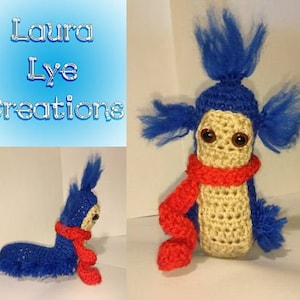 May include: A crocheted blue and tan creature with a red scarf. The creature has a long, thin body and a fluffy blue head. The creature is wearing a red scarf around its neck. The creature is a handmade toy.