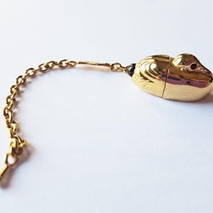 Puede incluir: Una cadena dorada con un pequeño colgante dorado con forma de pato. El pato tiene un ojo de piedra preciosa roja.