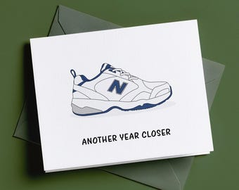 May include: A white greeting card featuring a graphic illustration of a white and navy blue athletic trainer with a grey heel detail. The text "ANOTHER YEAR CLOSER" is printed in black capital letters below the trainer illustration.