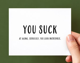 Puede incluir: Una tarjeta de felicitación rectangular blanca con texto en negro que dice YOU SUCK AT AGING. SERIOUSLY, YOU LOOK INCREDIBLE. La tarjeta presenta un diseño limpio con letras sans-serif centradas en la parte frontal.