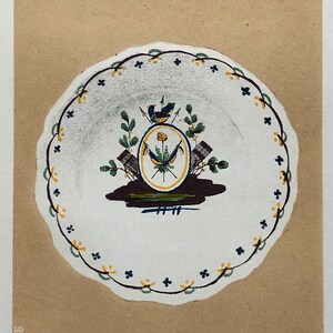 May include: A white ceramic plate with a blue, yellow and green floral design and a bird. The plate has a scalloped edge and a blue and yellow border. The text "ASSIETTE PATRIOTIQUE 1789" is printed below the plate.