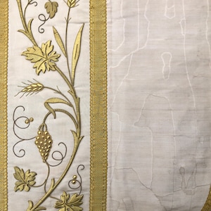 Puede incluir: Un panel de tela blanca con bordado dorado de una vid con hojas, uvas y espigas de trigo. La tela está bordeada con un ribete dorado.