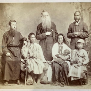 May include: A black and white photograph of seven people, including a man with a long beard wearing a dark robe, a woman wearing a long dress and a headscarf, and a young boy wearing a hat and a striped shirt. The group is posed in a studio setting with a backdrop of a wall and a fern.