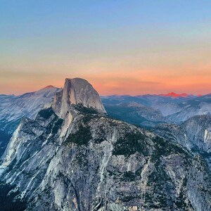 May include: A panoramic view of Half Dome, a granite rock formation in Yosemite National Park, California. The sky is a vibrant pink and orange at sunset, with the mountains in the foreground appearing blue and gray.