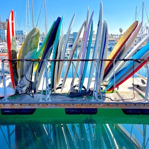 May include: A rack of colorful surfboards leaning against a wooden dock, with a blue sky and boats in the background.