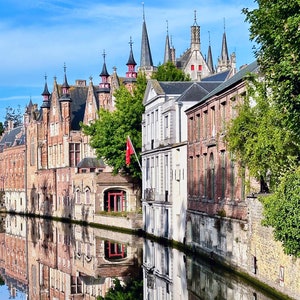 May include: A view of a canal in Bruges, Belgium, with traditional brick buildings and a reflection of the buildings in the water. The buildings have red tile roofs and some have spires. A red flag is hanging from one of the buildings.