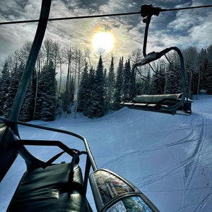 May include: A ski lift ascends a snow-covered slope on a bright, sunny day. The lift chairs are empty, and the sun is partially obscured by a bright halo. Tall evergreen trees line the sides of the ski run, and the sky is a mix of blue and gray.