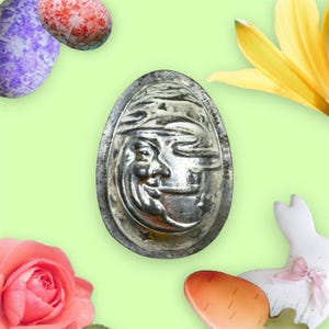 May include: An antique silver metal Easter egg mould featuring a smiling moon face design. The mould is surrounded by colourful Easter eggs, a pink rose, a yellow flower, and a white bunny with a pink bow on a light green background.